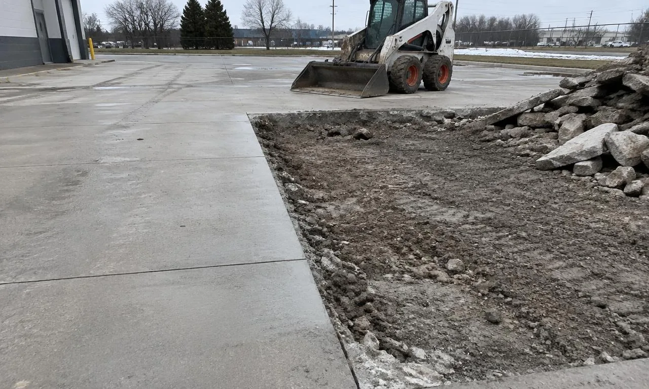 Cut concrete slab beside removed section on commercial lot with skid steer in background