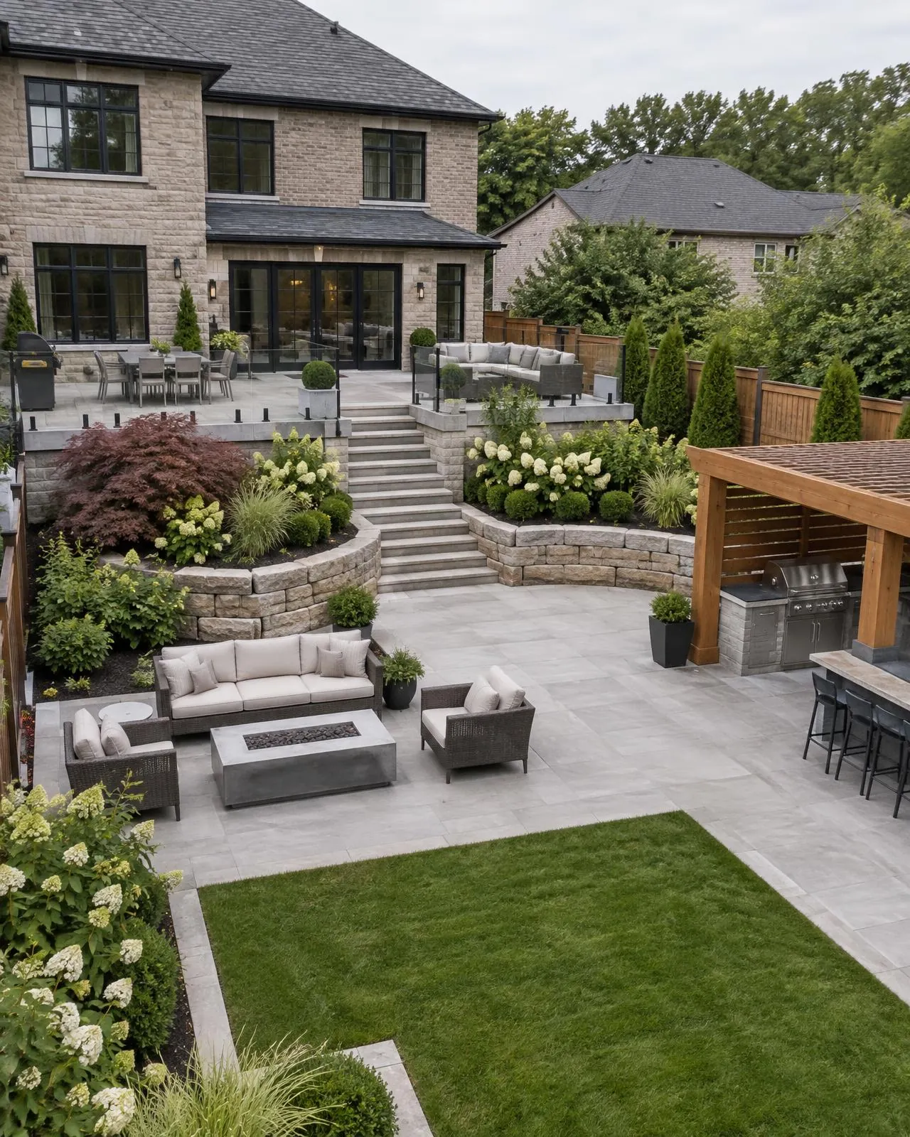 Multi-level backyard with terraces, stone steps, retaining wall, outdoor kitchen, fire table and planting behind a stone home