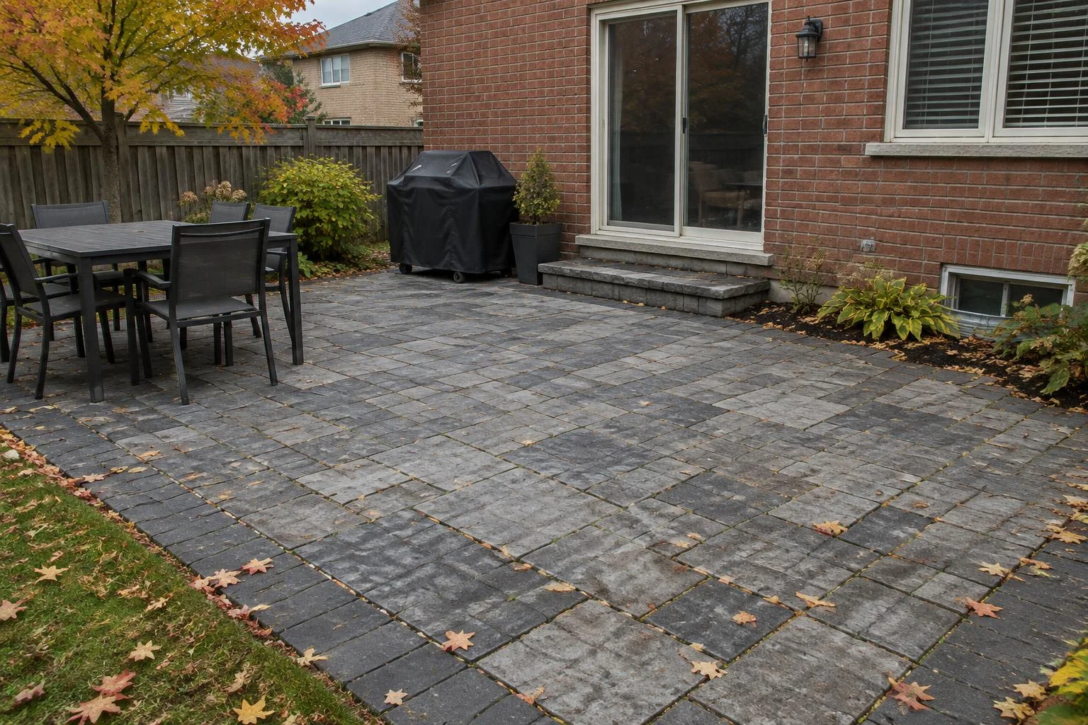 Grey and charcoal paver patio with light wear and autumn leaves behind a brick house