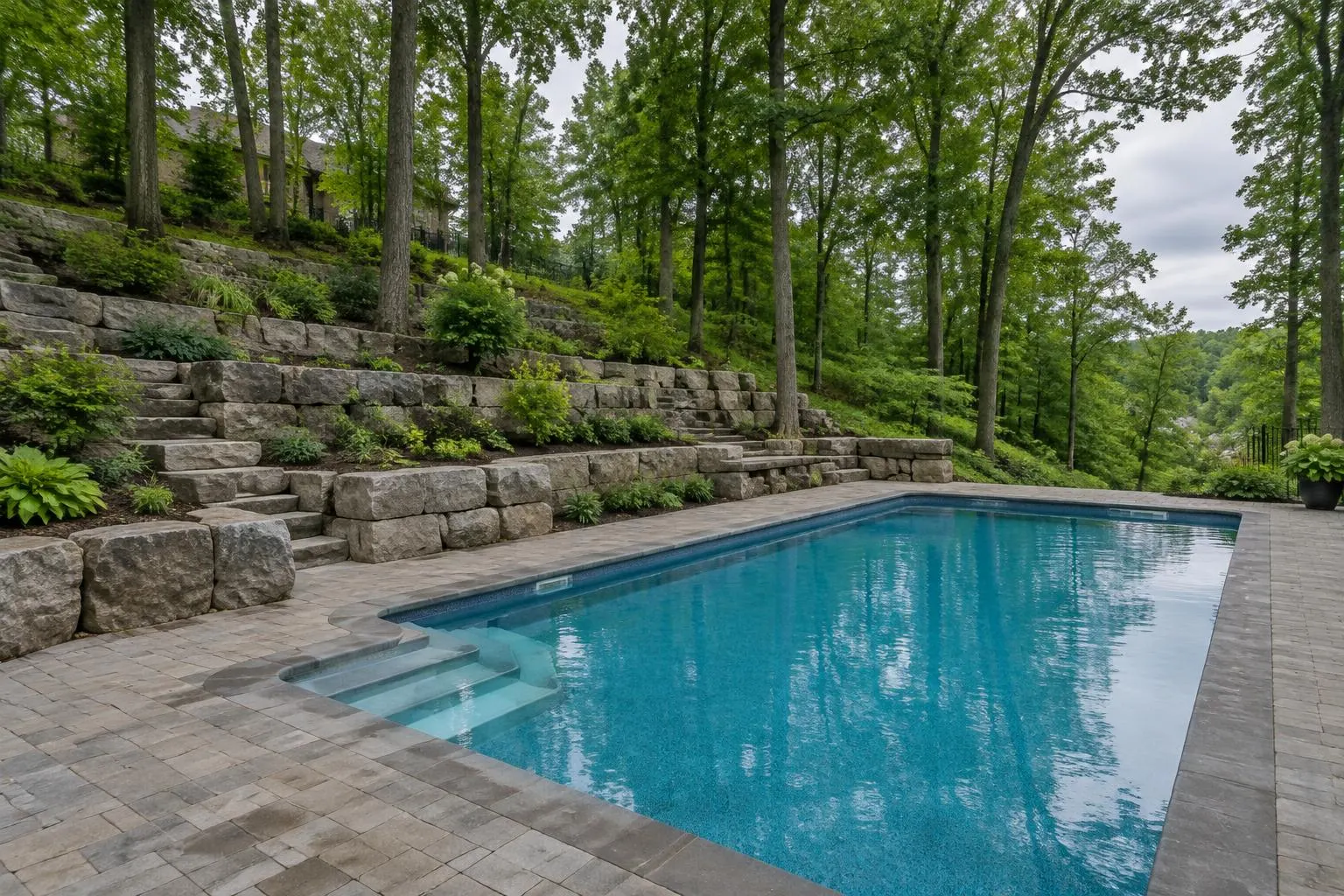 Inground pool on a sloped wooded lot with tiers of armour-stone retaining walls stepping up the hillside