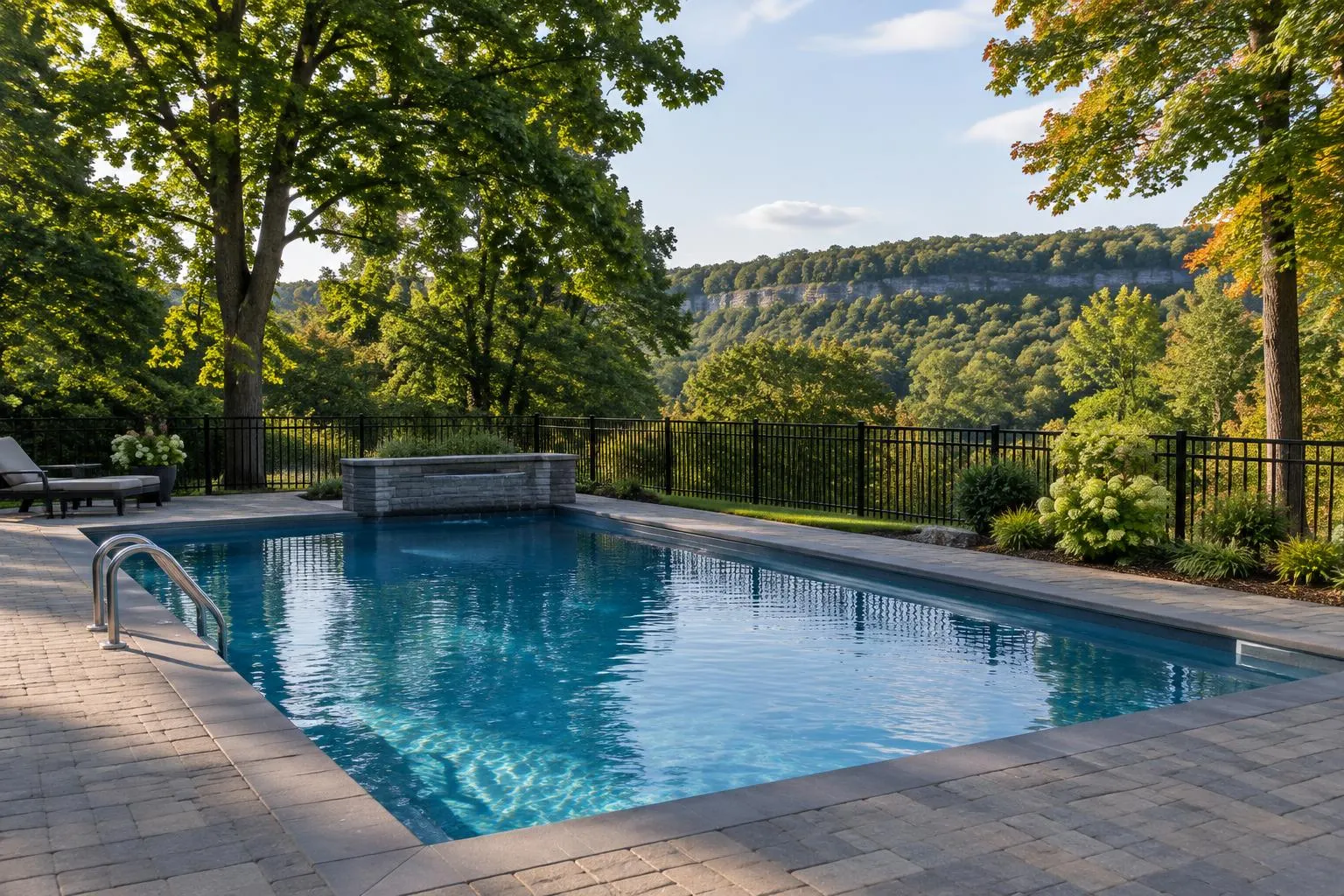 Inground pool with a paver deck and black aluminum fence, a wooded escarpment ridge and mature maples behind it