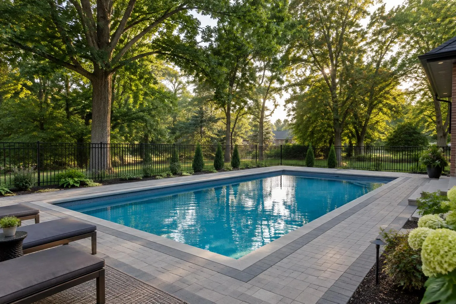 Rectangular inground pool with a grey paver deck and black aluminum fence under mature trees