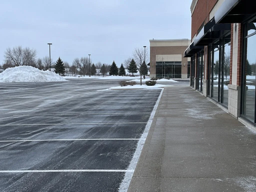 Plowed commercial plaza parking lot at dawn with cleared, salted walkways in front of the storefronts